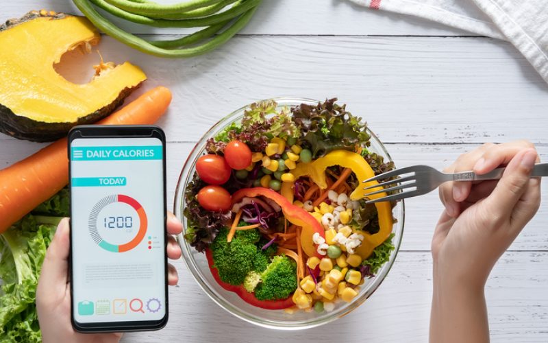 Calories counting , diet , food control and weight loss concept. Calorie counter application on smartphone screen at dining table with salad, fruit juice, bread and fresh vegetable. healthy eating